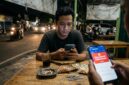 Ilustrasi: Seorang pria cemas memeriksa ponselnya saat kehabisan pulsa di sebuah warkop malam hari di pinggir jalan Jakarta.(poto: AI).